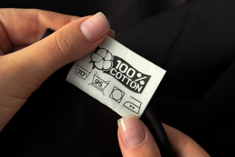 Dłonie trzymające metkę ubrania z oznaczeniem 100% bawełny i symbolami prania