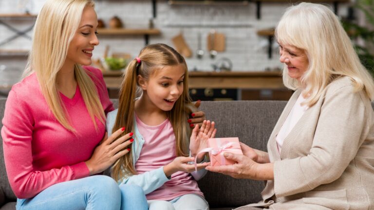 Babcia wręcza wnuczce prezent w różowym pudełku, obok siedzi mama